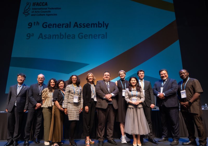 A group of people standing on a stage. They are the new Board of the International Federation of Arts Councils and Cultural Agencies: Hailin Yu, Adrian Collette AM, Michelle Chawla, Magdalena Moreno Mujica, Julie Diphofa, Kristin Danielsen, Humberto López La Bella, Kajsa Ravin, Nguyen Phuong Hoa, Rafael Ivorra Zaragoza, Emile Kairua and Maanka Chipindi.